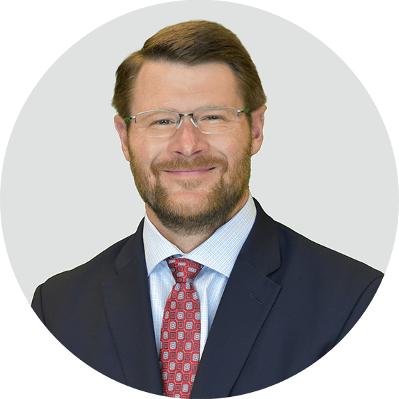 A man with short brown hair, glasses, and a beard is wearing a dark suit, light blue shirt, and red patterned tie, smiling at the camera against a light gray background.