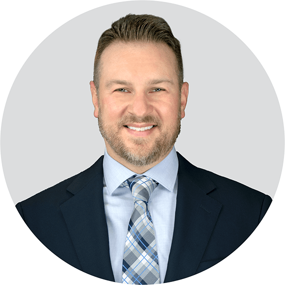 A man with short brown hair and a trimmed beard wearing a dark suit, light blue shirt, and a blue plaid tie, smiling at the camera against a light gray background.