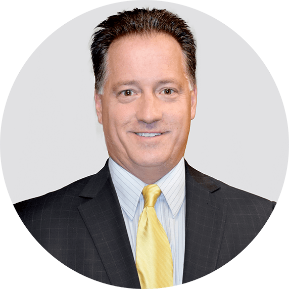 A man with short dark hair, wearing a black pinstripe suit, light blue shirt, and yellow tie, smiles at the camera against a light gray circular background.