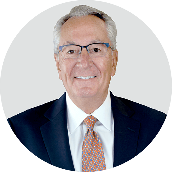A smiling older man with gray hair and glasses, wearing a dark suit, white shirt, and patterned tie, posed against a plain light gray background.