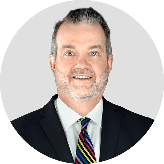 A middle-aged man with light skin, gray hair, and a beard, wearing a black suit, white shirt, and a multicolored striped tie, smiling in front of a plain light gray background.