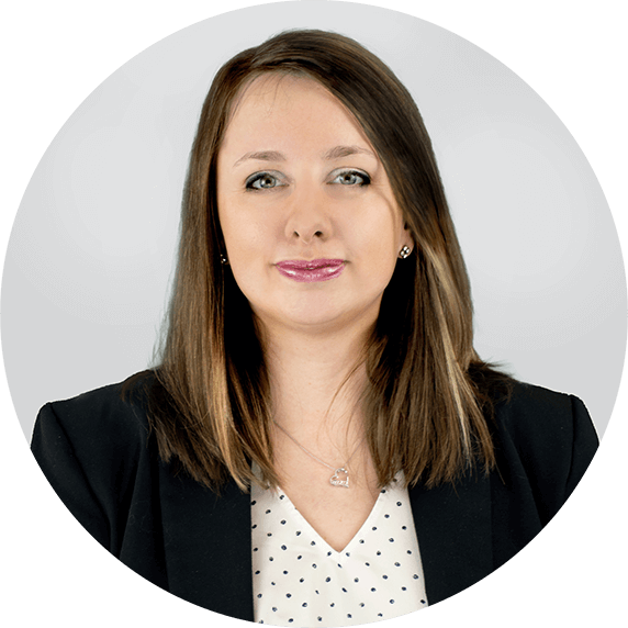 Woman with straight, shoulder-length brown hair wearing a black blazer over a white blouse with small black dots, posing against a light gray background.