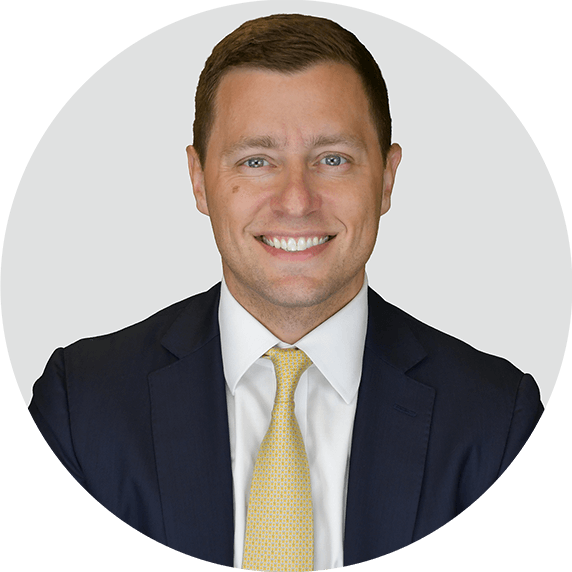 Smiling man with short brown hair wearing a dark suit, white shirt, and yellow tie, posing against a light gray background.