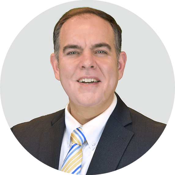 A man in a dark suit, white shirt, and striped yellow and blue tie smiles at the camera against a light gray circular background.