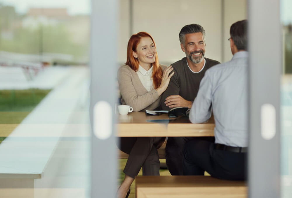 A smiling couple sits at a table across from a man in an office setting, having a friendly discussion. The woman has her hand on the mans shoulder, and they all appear engaged and relaxed.