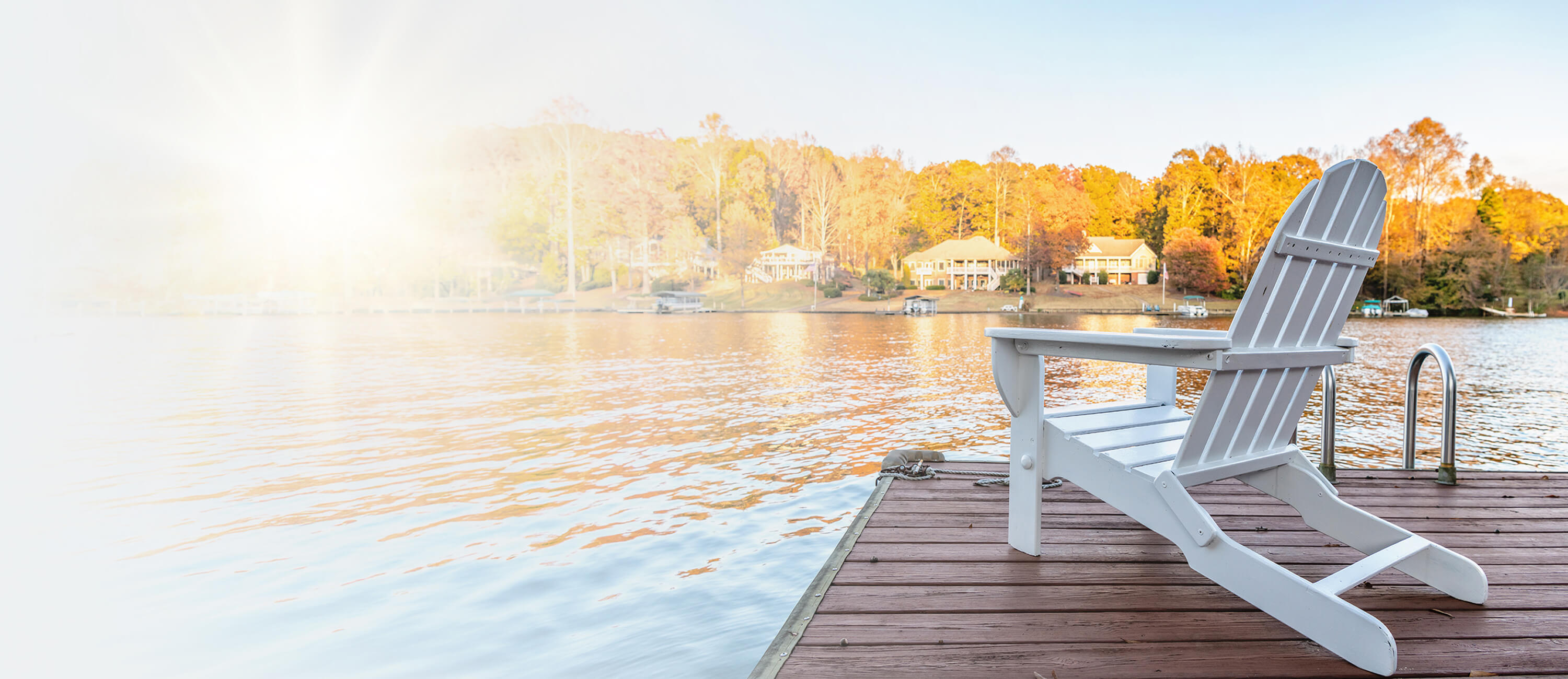 A white Adirondack chair sits on a wooden dock overlooking a calm lake, with trees in autumn colors and houses in the background under a bright, sunny sky.