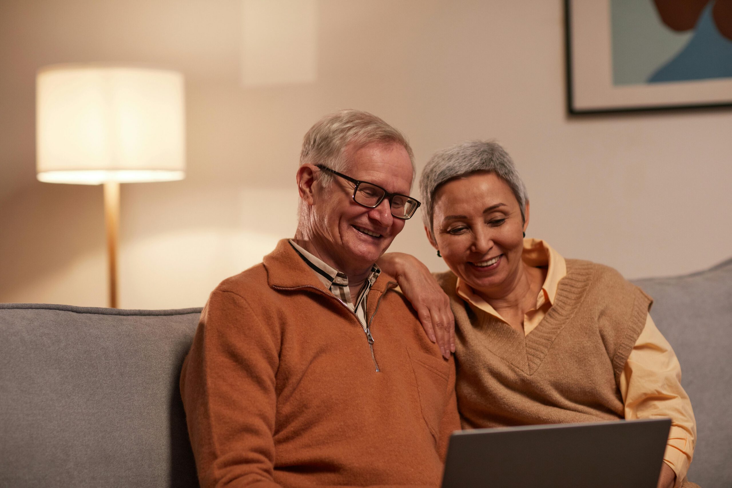 An older couple sits closely together on a sofa, smiling as they look at a laptop screen. The room is warmly lit with a lamp in the background, creating a cozy atmosphere.