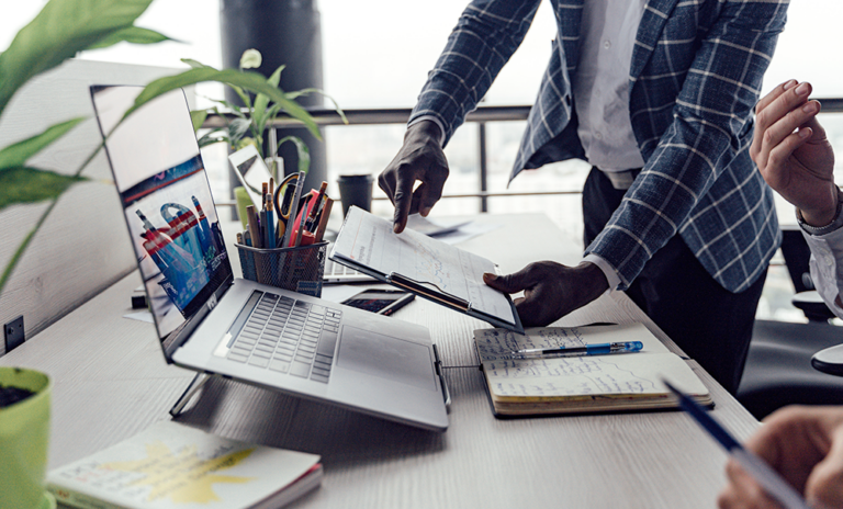Two people work at a desk with a laptop, notebooks, and office supplies. One person is standing and pointing to a document, while the other person is seated, holding a pen. The workspace has a modern, collaborative feel.