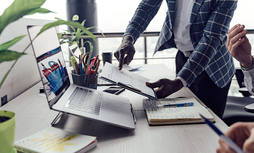 Two people work at a desk with a laptop, notebooks, and office supplies. One person is standing and pointing to a document, while the other person is seated, holding a pen. The workspace has a modern, collaborative feel.