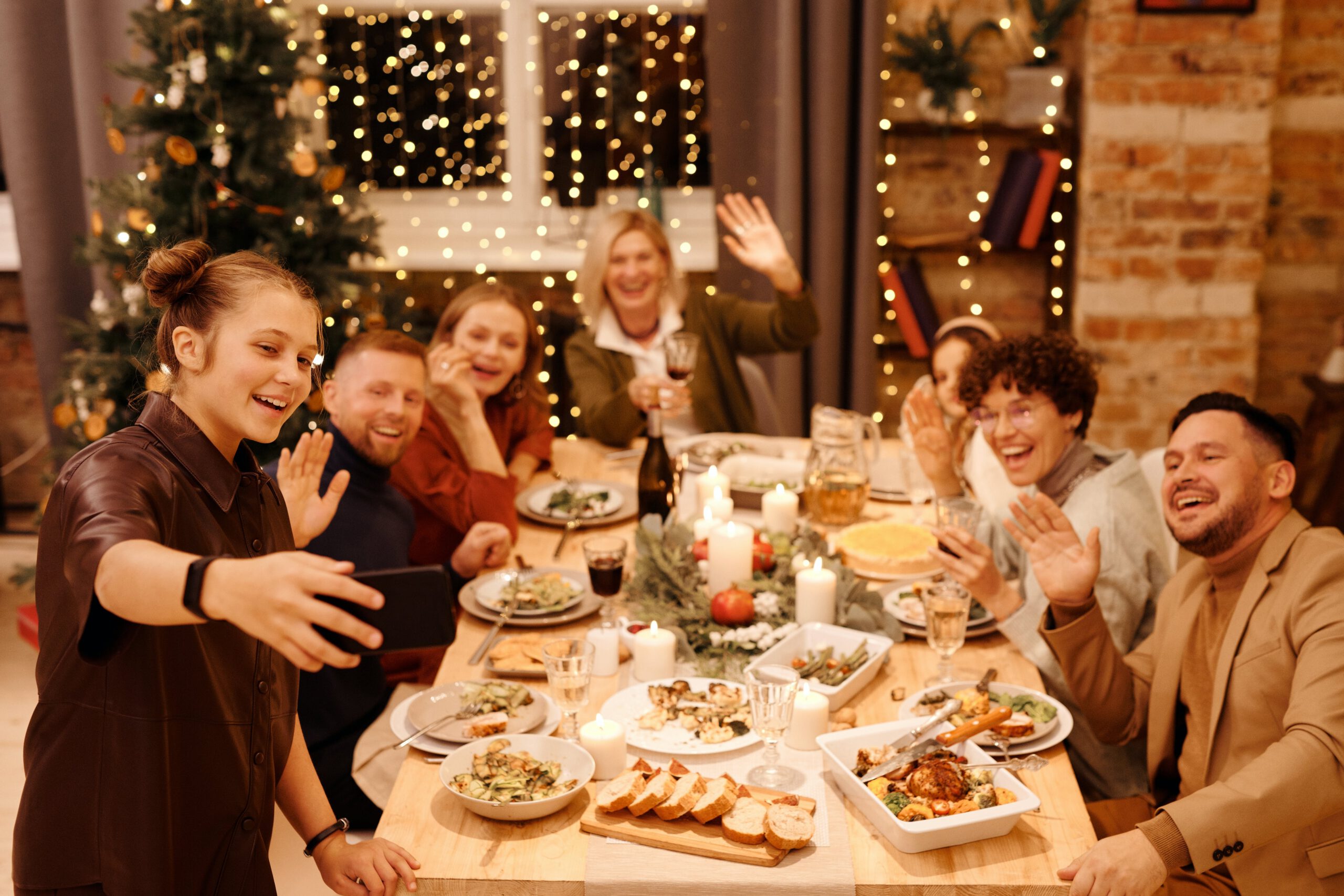 A group of people smiling and waving at the camera while taking a selfie at a festive dinner table, decorated with candles and food, with a Christmas tree and string lights in the background.