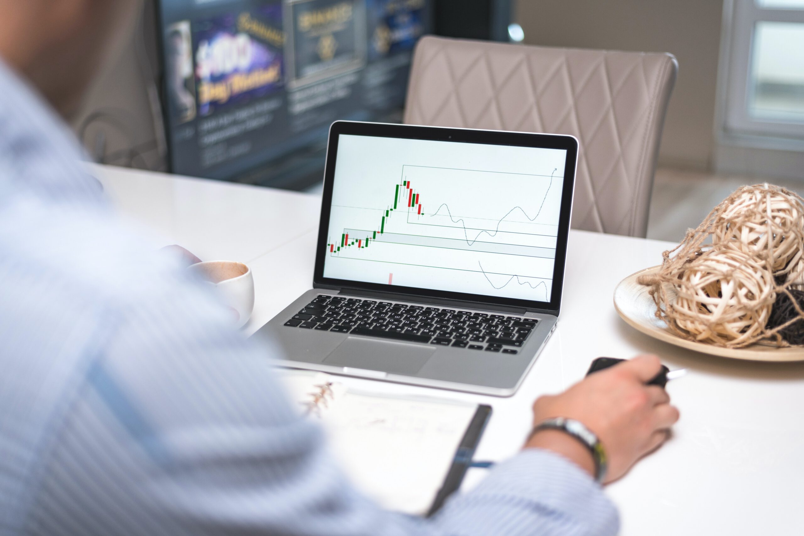 A person analyzes a candlestick chart with trend lines on a laptop at a desk, holding a computer mouse. A notebook, coffee cup, and decorative bowl are also on the table.