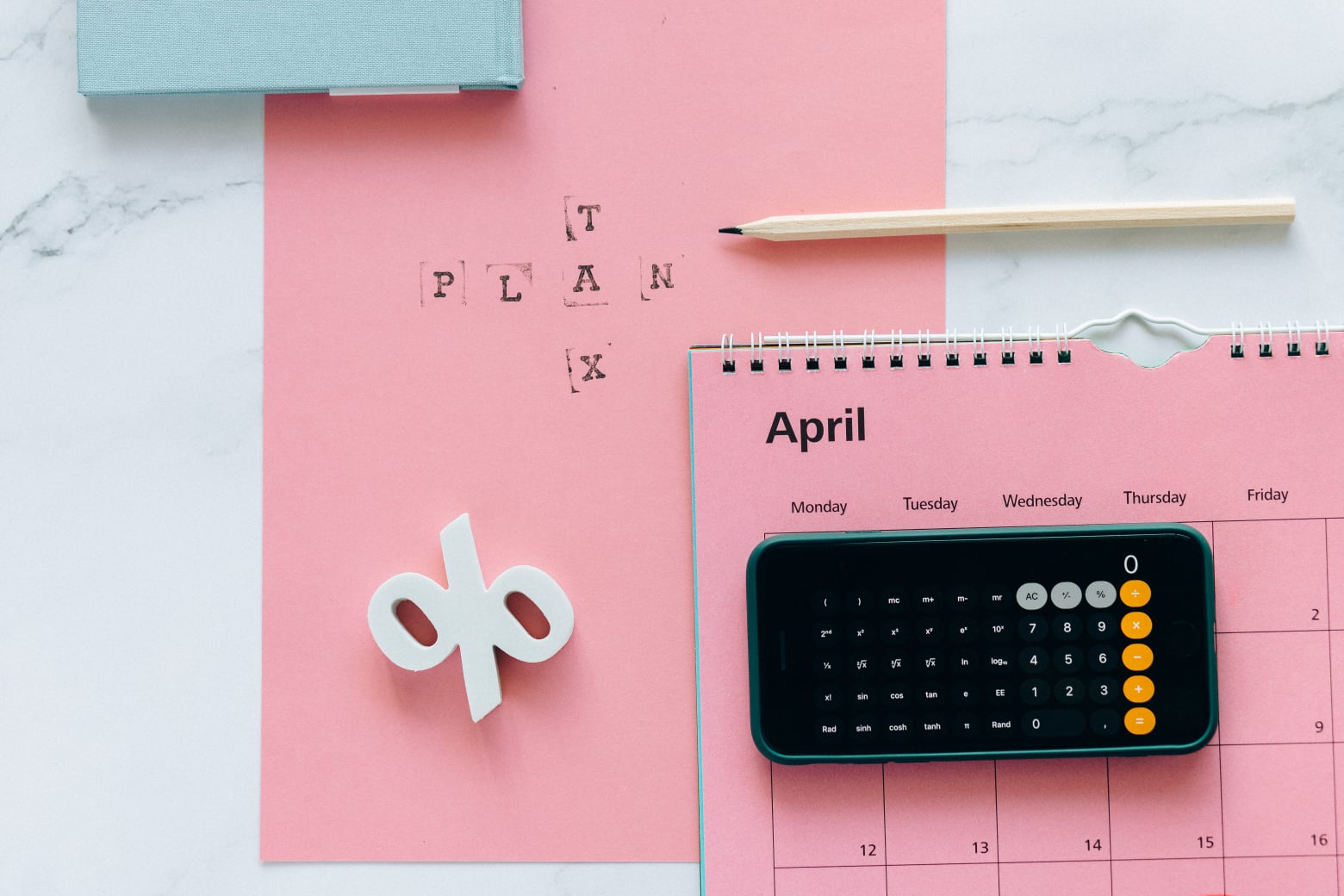 A pink calendar open to April, a smartphone with a calculator app, a pencil, a book, and a paper with PLAN stamped on it, arranged on a white desk.