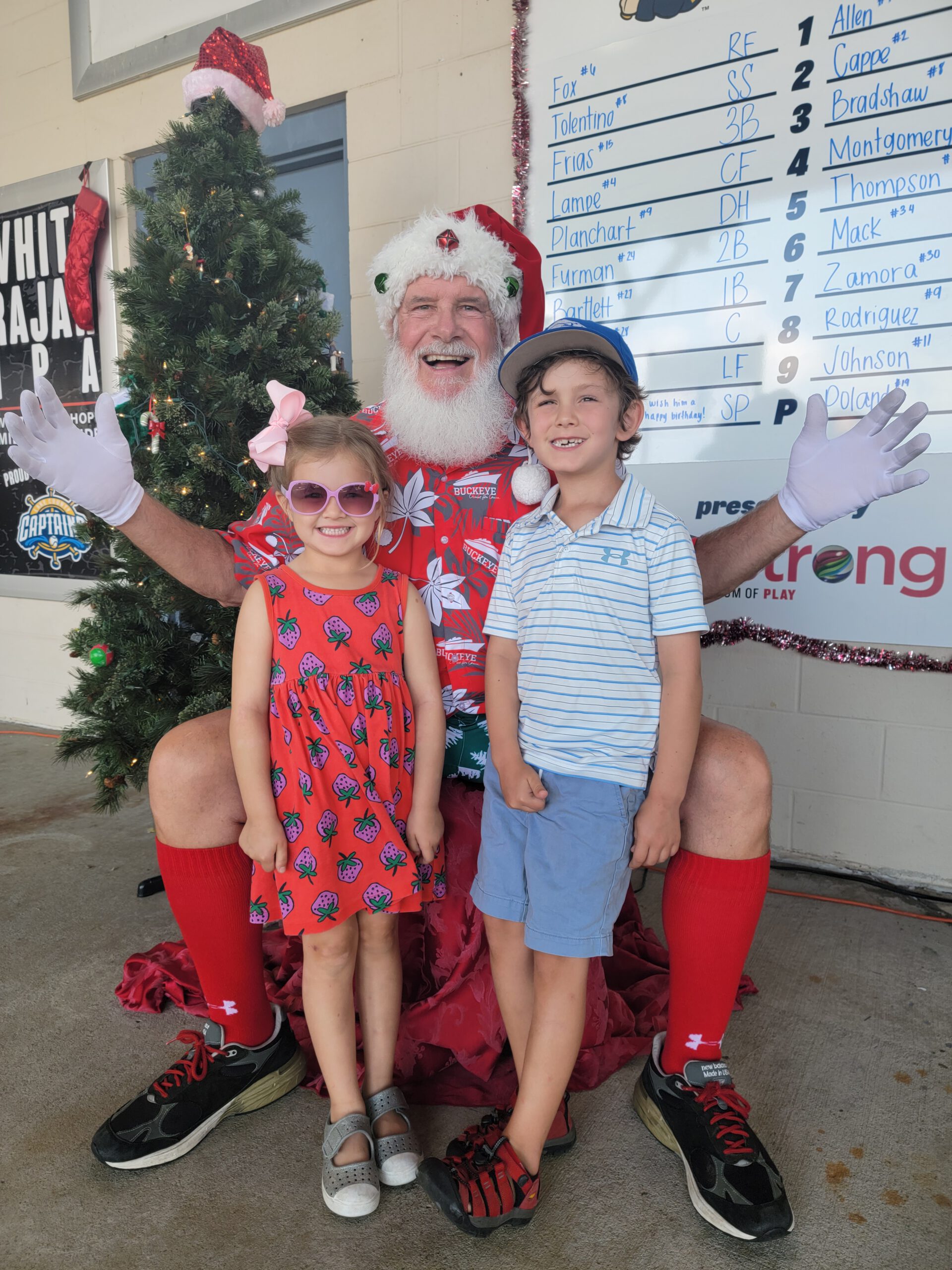 Santa Claus in a Hawaiian shirt and red shorts poses with a smiling young boy and girl in summer clothes, standing by a decorated Christmas tree and a baseball lineup board.