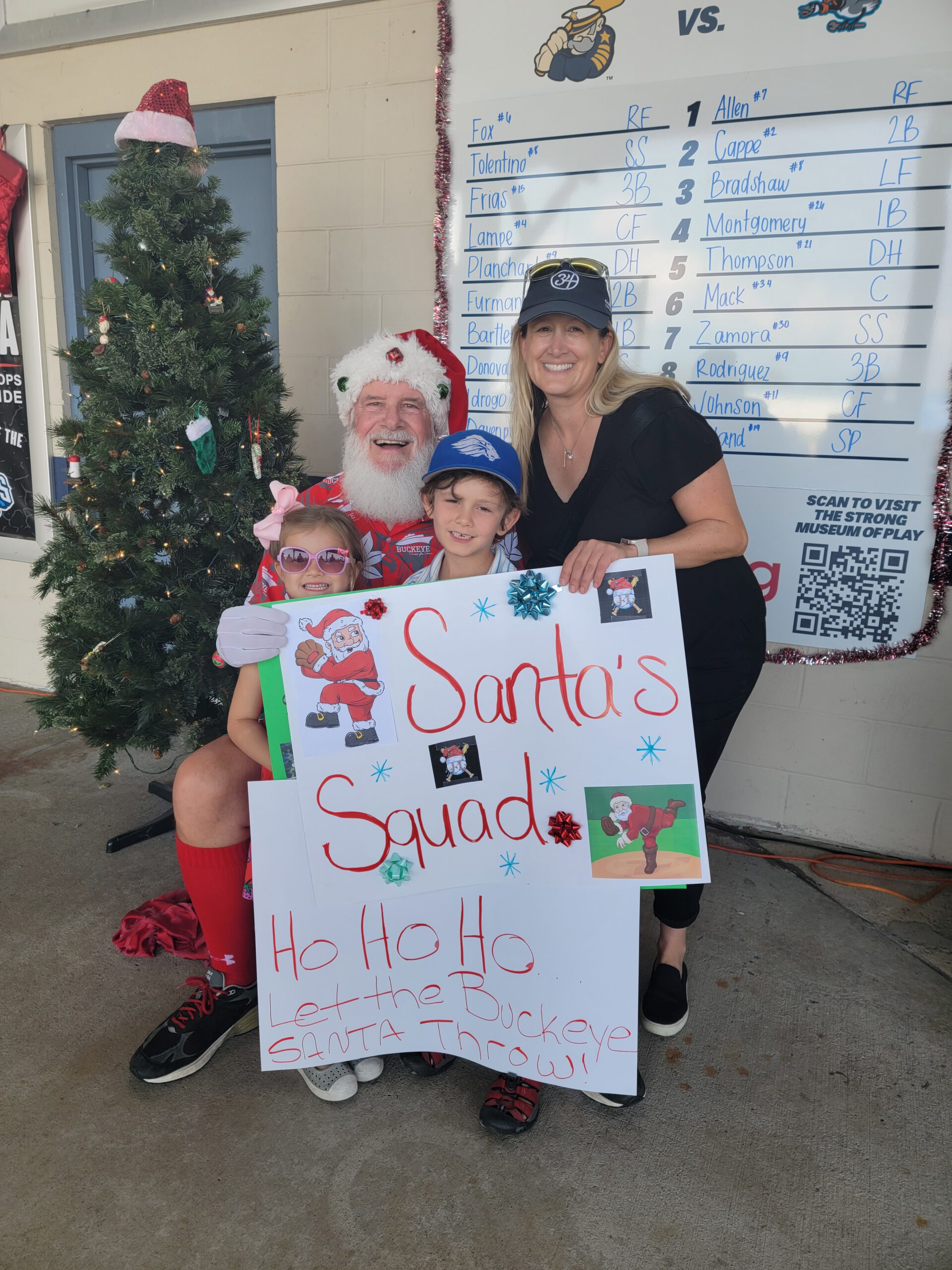 A man dressed as Santa Claus sits with two children and a woman in front of a Christmas tree, holding a festive sign that reads Santas Squad—Ho Ho Ho Let the Buckeye Santa Throw! at a sports event.