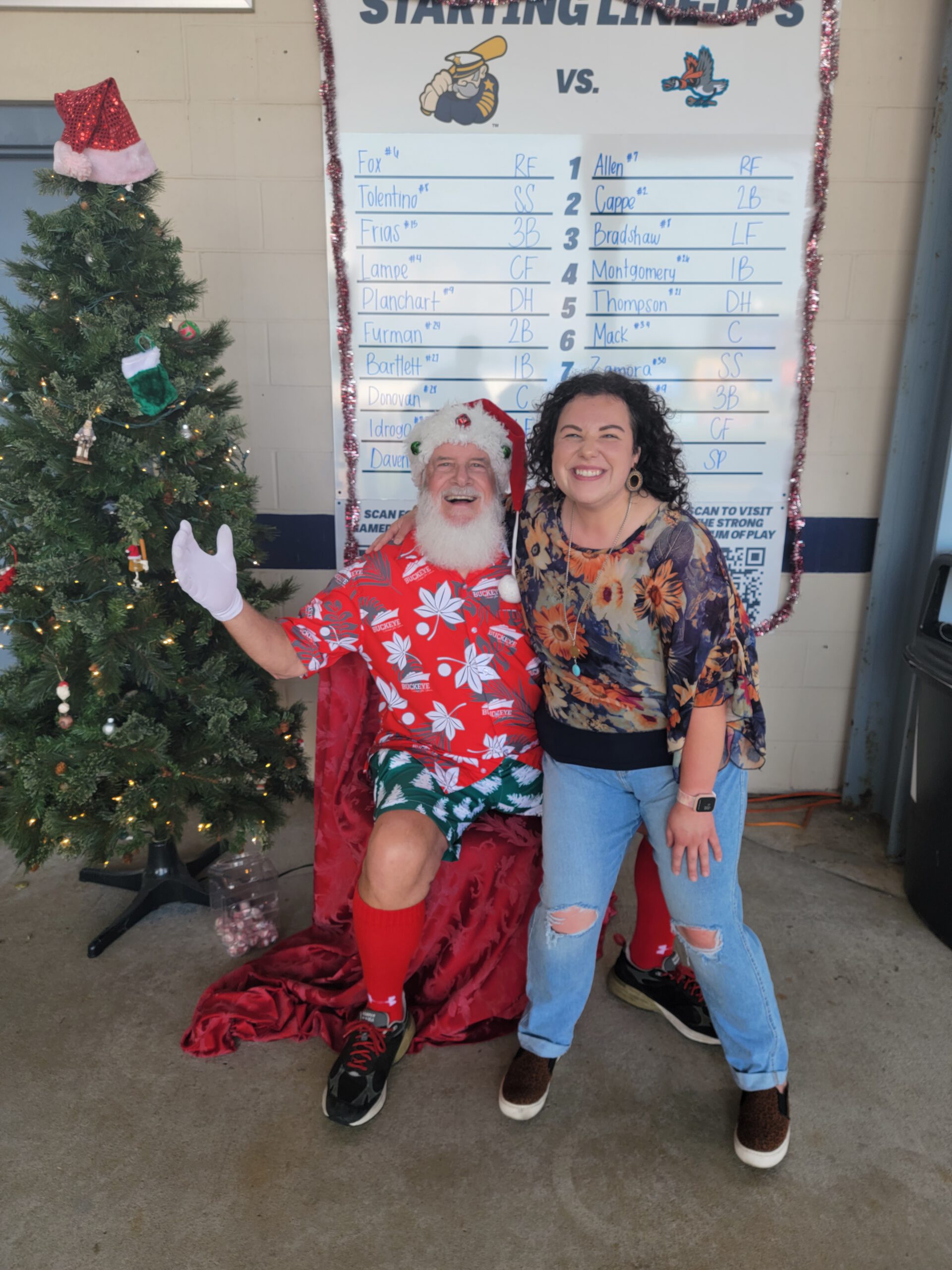 A man dressed as a Hawaiian Santa poses next to a decorated Christmas tree, smiling with a woman in casual clothes. Behind them is a large board displaying a baseball teams starting lineup.