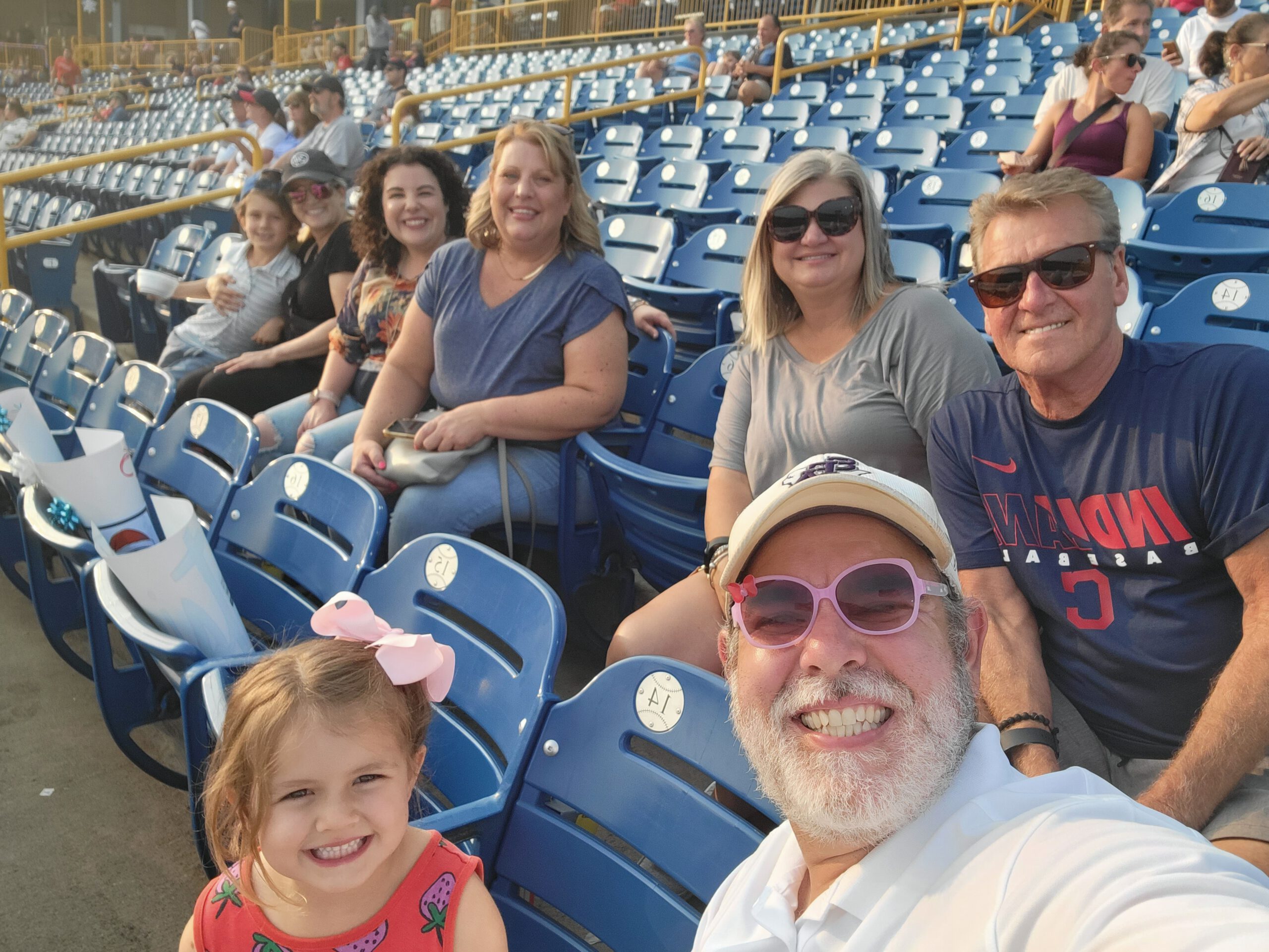 A group of smiling people, including young children and adults, sit together in blue stadium seats, enjoying a sunny day at a baseball game. The stands around them have many empty seats.