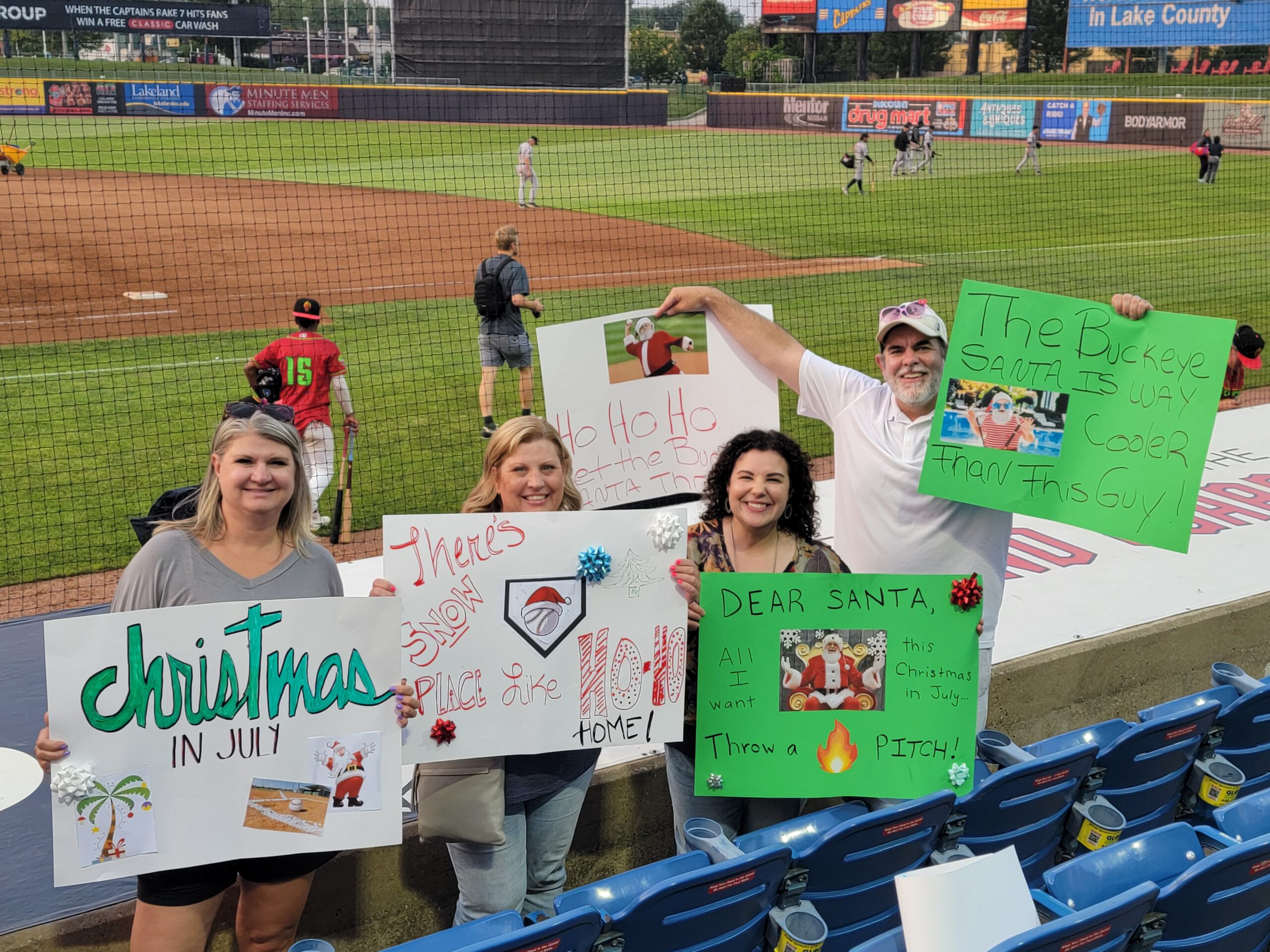 Four smiling people hold colorful, festive signs with Christmas themes at a baseball stadium. Players are on the field in the background, and the stands are mostly empty. The signs read messages like “Christmas in July” and “Dear Santa.”.