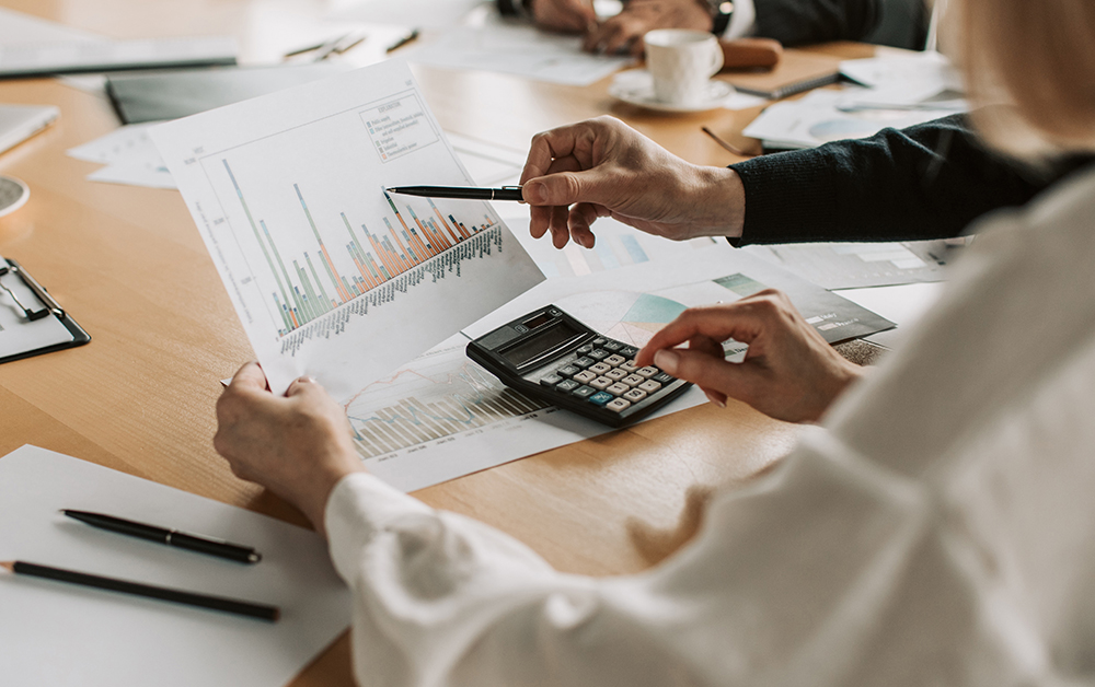 Two people analyze a printed bar graph at a desk, one pointing at the chart with a pen while the other uses a calculator. Papers, pens, and a coffee cup are scattered on the table.