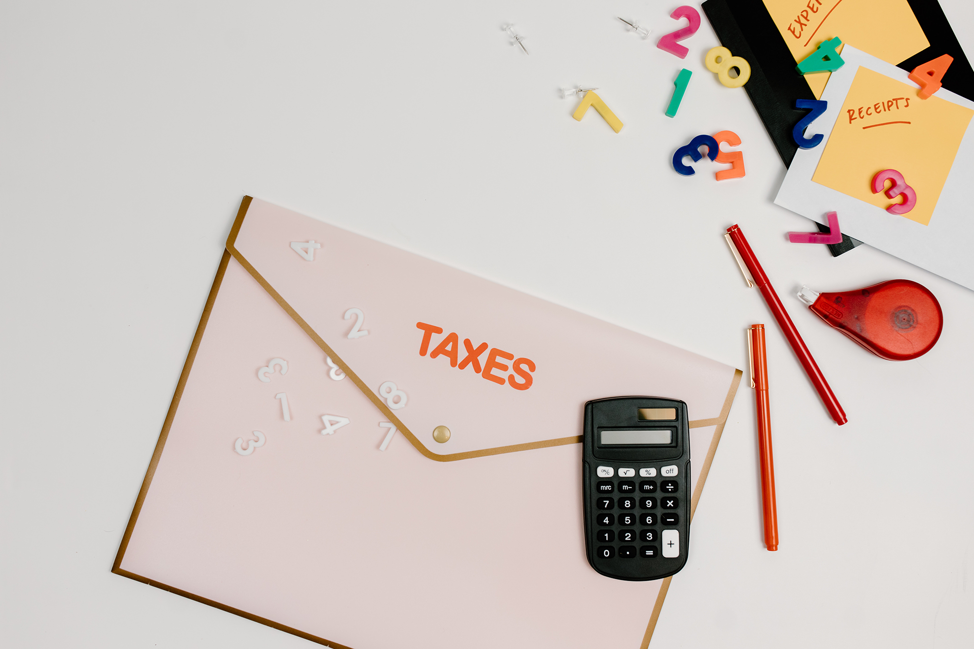 A pink folder labeled TAXES, a calculator, red pen, tape dispenser, and scattered number magnets on a white desk with organized receipts and expense folders.