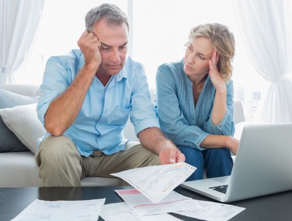 A worried middle-aged couple sits on a couch, looking at bills and paperwork on a table in front of them, with a laptop open, appearing stressed or concerned about finances.