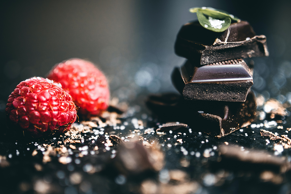 Two fresh raspberries sit next to a small stack of dark chocolate pieces, sprinkled with chocolate shavings, on a dark surface. A green mint leaf and sea salt garnish the chocolate.