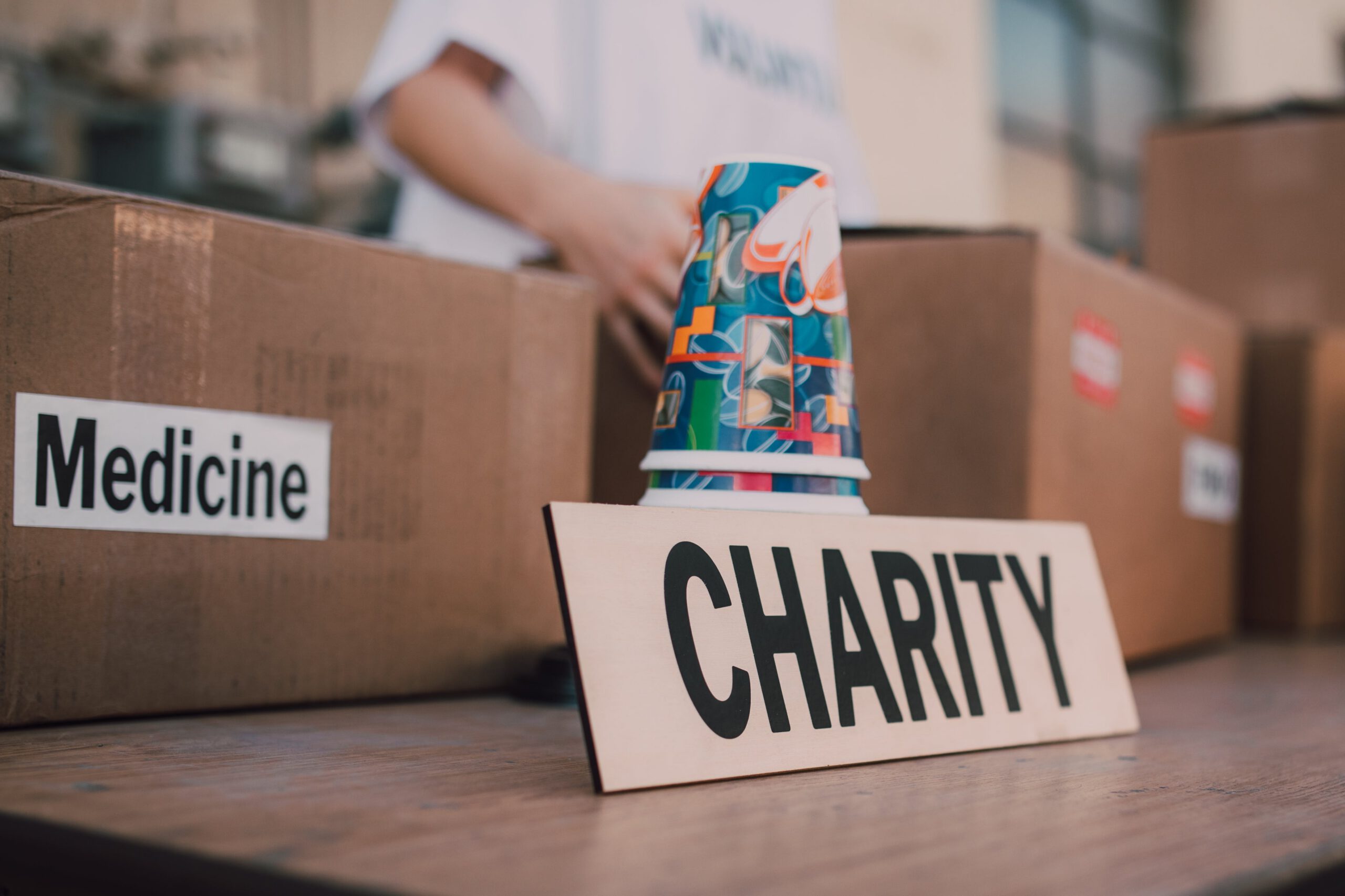 A table with cardboard boxes labeled Medicine and a sign reading Charity. A colorful paper cup and a person in the background suggest a donation or charity event.