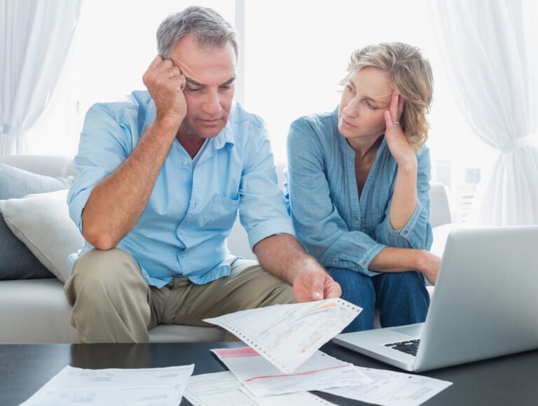 A middle-aged couple sits on a couch, looking stressed while reviewing bills and paperwork with a laptop on the table in front of them.