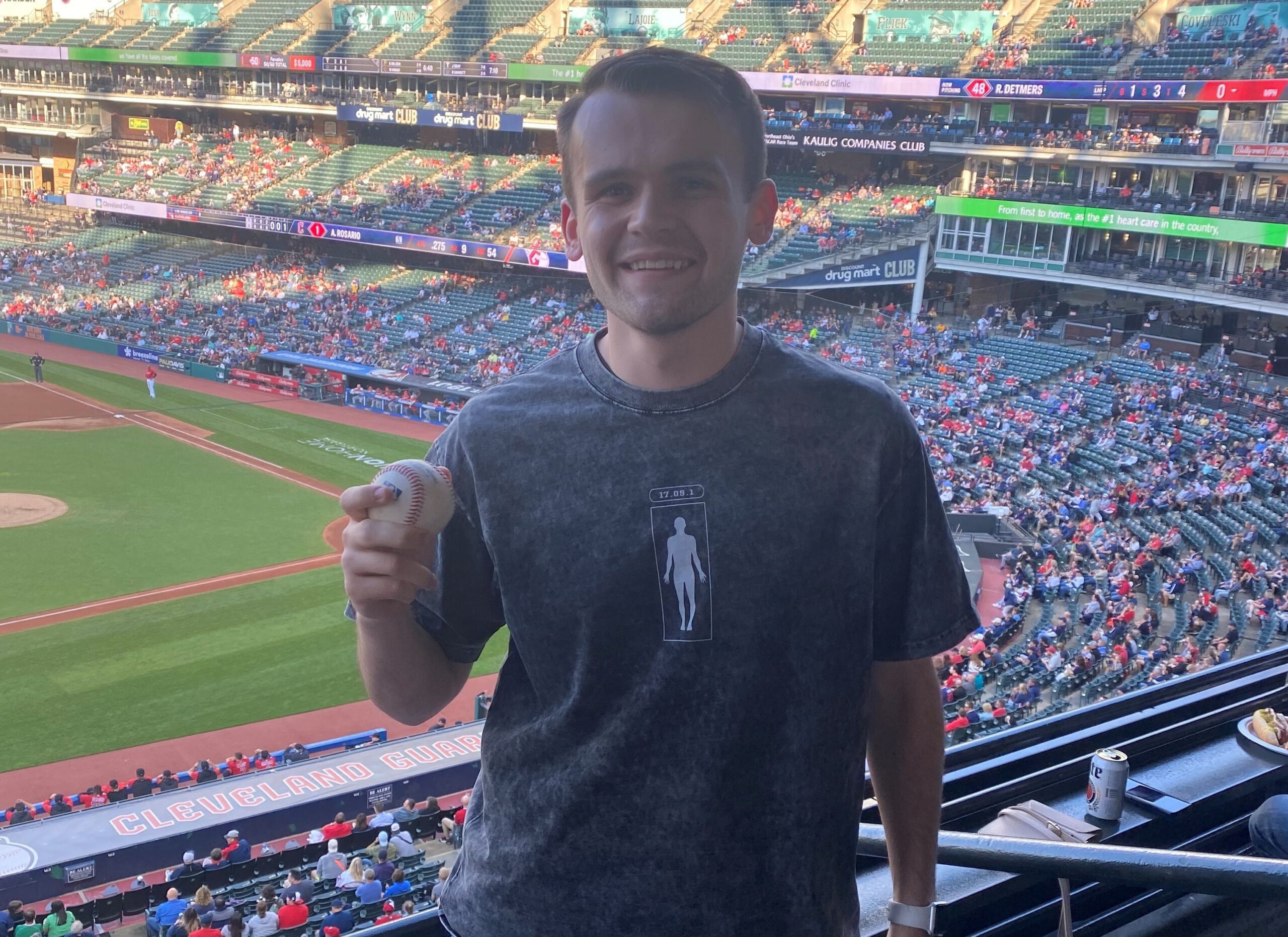A young man smiling and holding a baseball inside a stadium with Cleveland Guardians visible in the background. The stands are partially filled, and a game is taking place on the field.