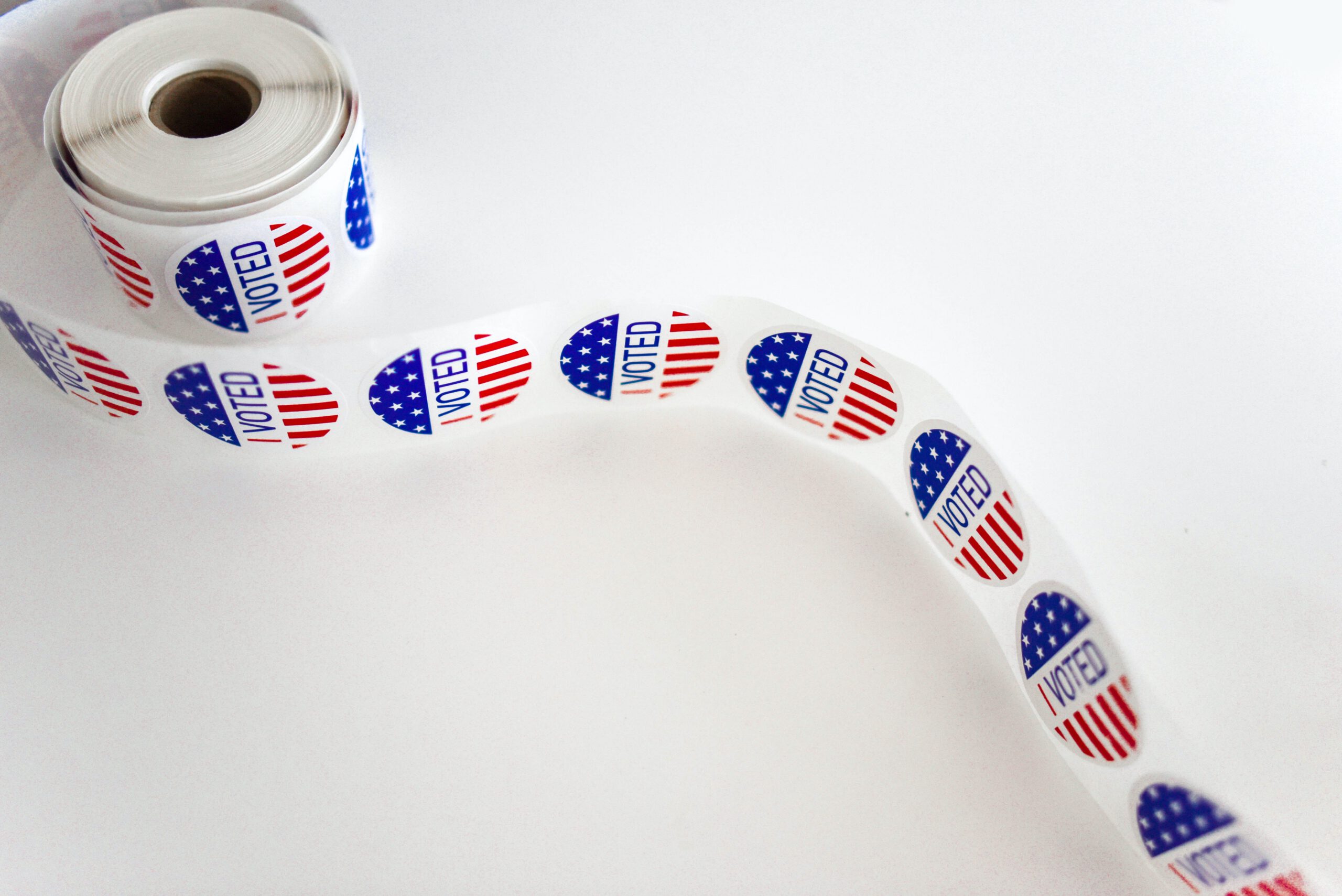 A roll of I Voted stickers featuring the U.S. flag design on a white background, with the stickers unspooled in a curve.