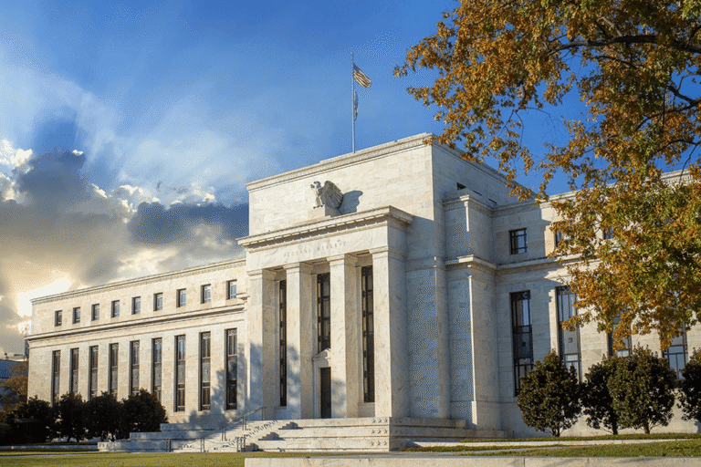 A large neoclassical white stone building with tall columns and an American flag on the roof, set against a dramatic sky with sunrays and surrounded by trees and greenery.