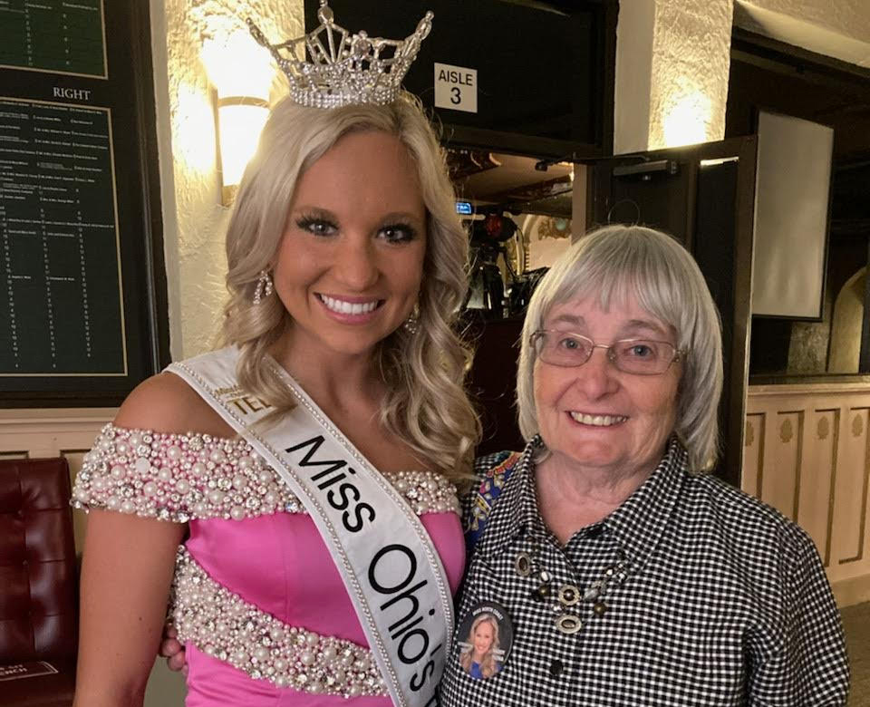 A young woman wearing a “Miss Ohio 2022” sash and crown stands smiling next to an older woman with gray hair and glasses, both posing indoors and looking at the camera.