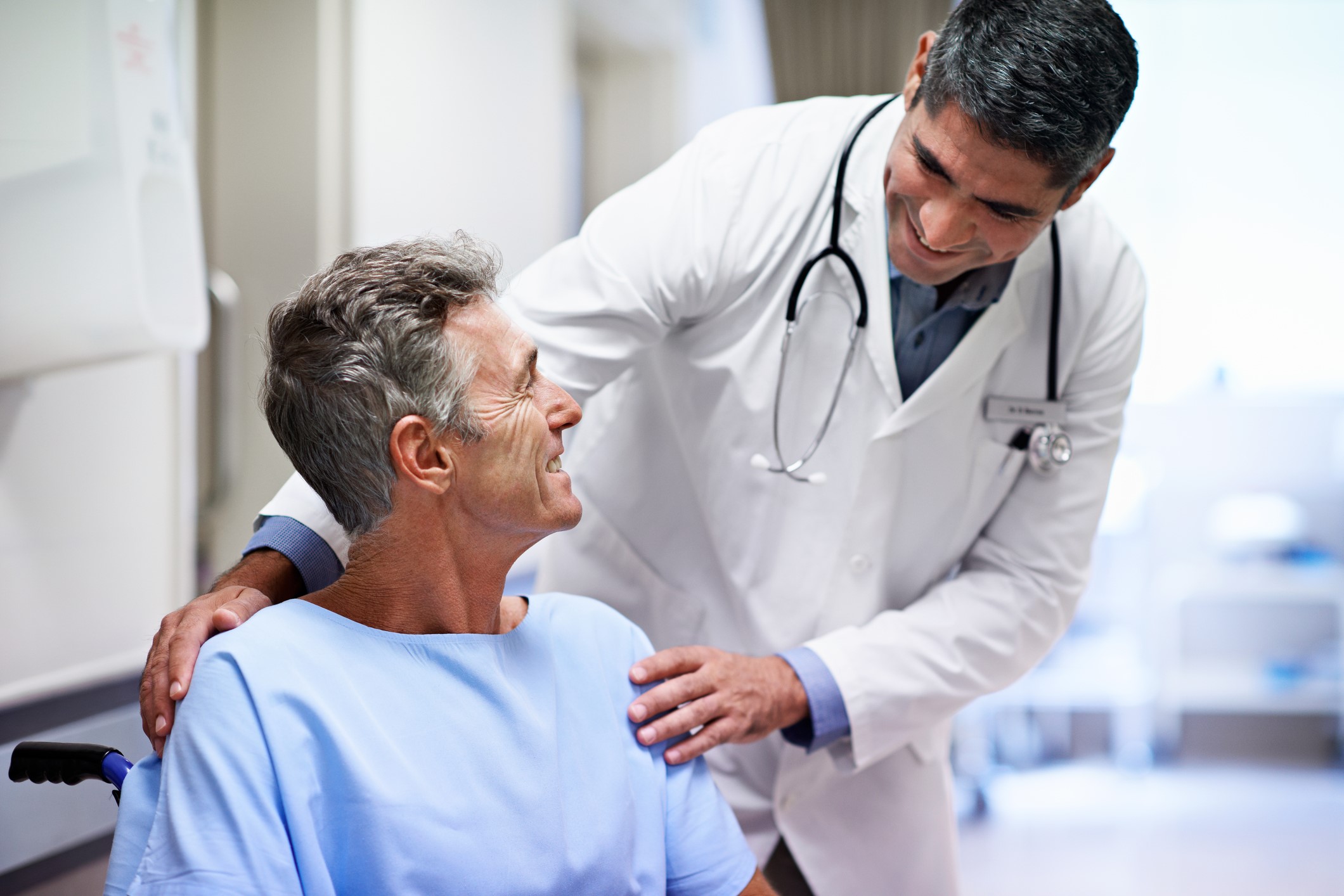 A doctor in a white coat with a stethoscope smiles and talks to an older male patient in a hospital gown, sitting in a wheelchair in a bright medical setting.