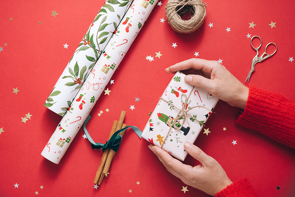 Hands holding a wrapped Christmas gift on a red surface with gold star confetti, decorative wrapping paper rolls, cinnamon sticks, twine, and scissors.