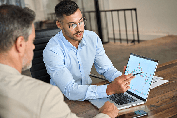 Two men sit at a desk, and one man points at a laptop screen displaying financial graphs and charts, while the other listens attentively. Papers and a phone are on the desk.