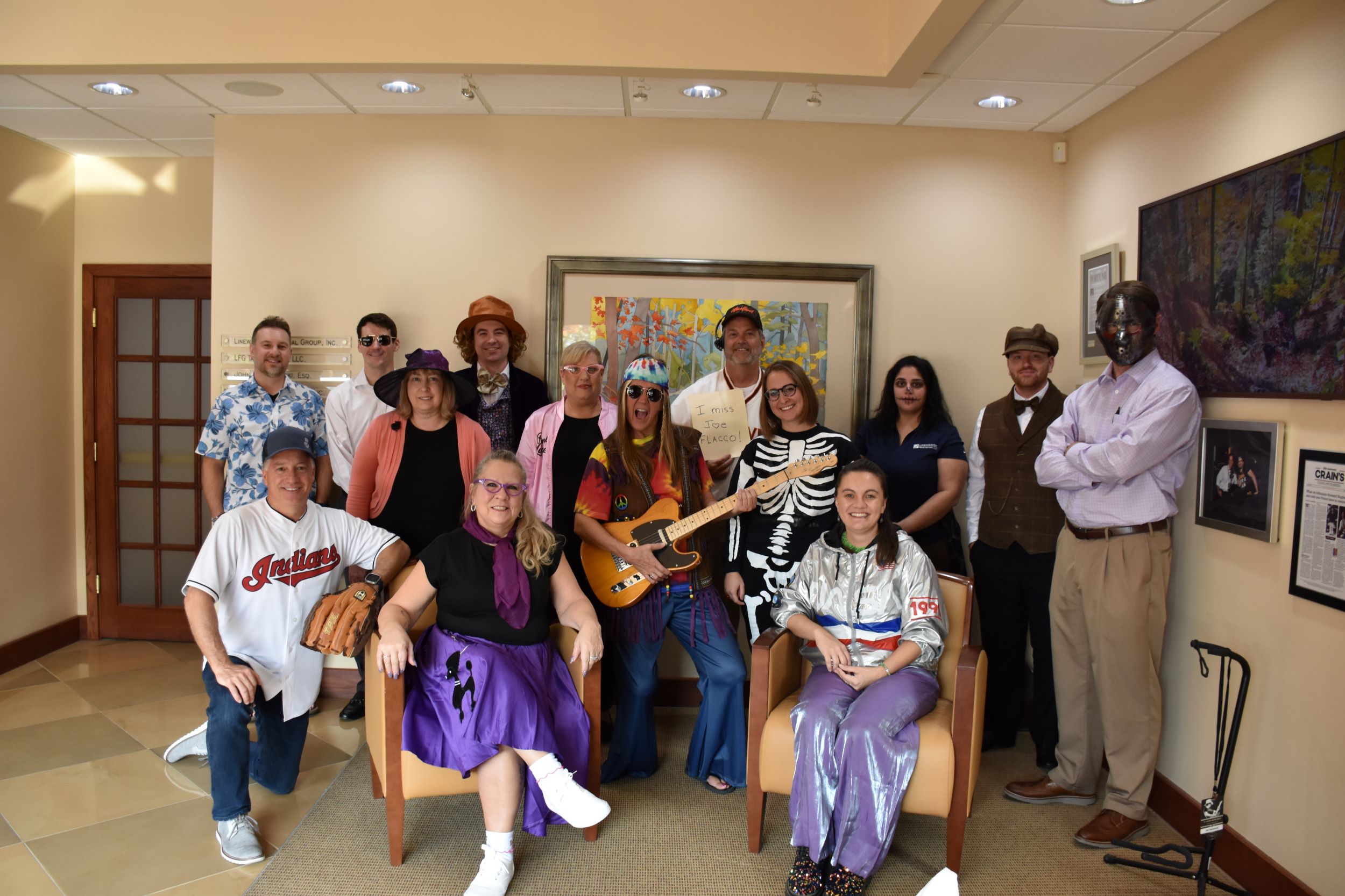 A group of people dressed in various costumes, including sports, retro, skeleton, and western outfits, pose together in an office space with artwork and framed photos on the walls.