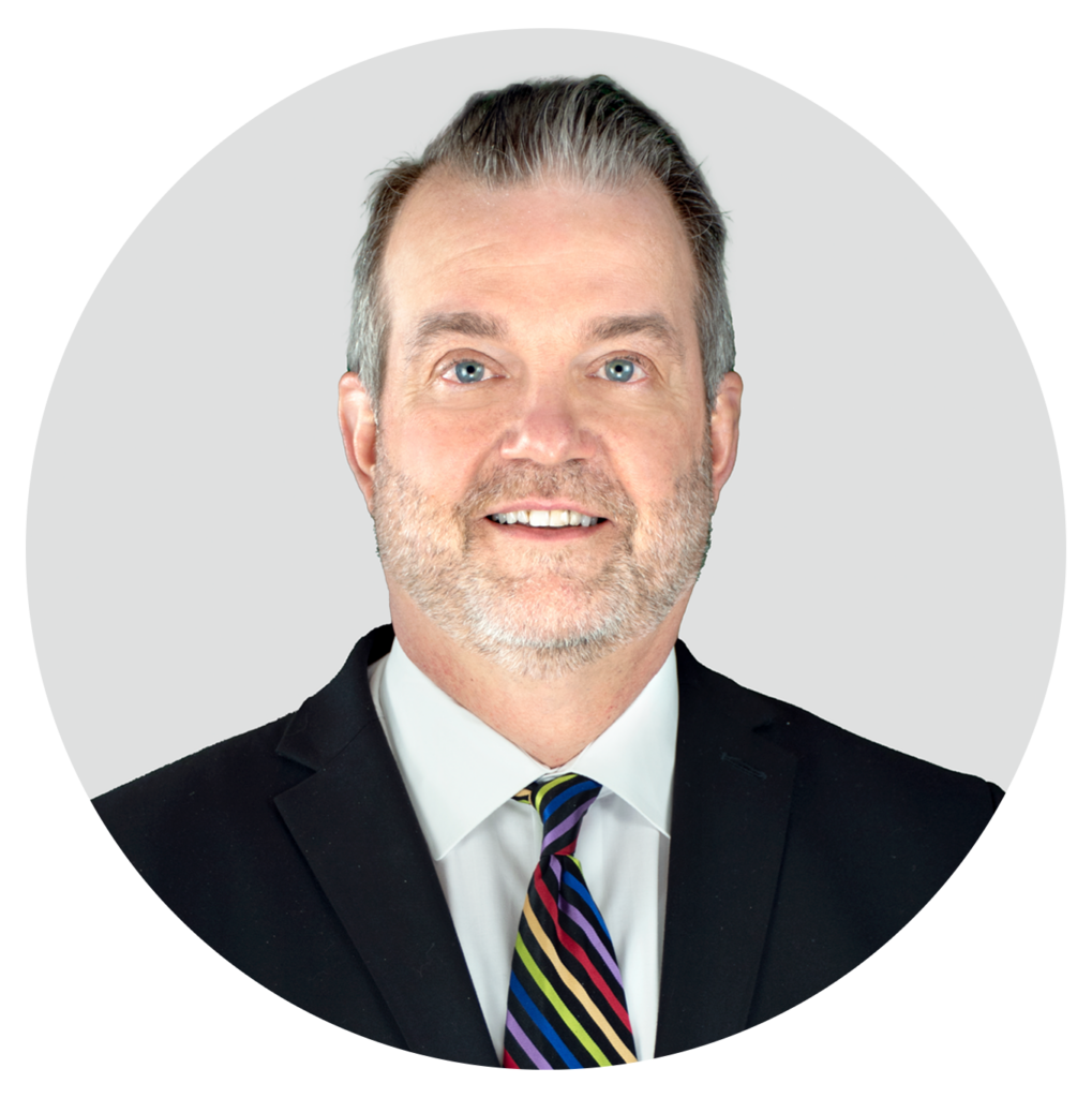 A man with short gray hair and a trimmed beard wearing a black suit, white shirt, and a colorful striped tie, smiling in front of a light gray background.