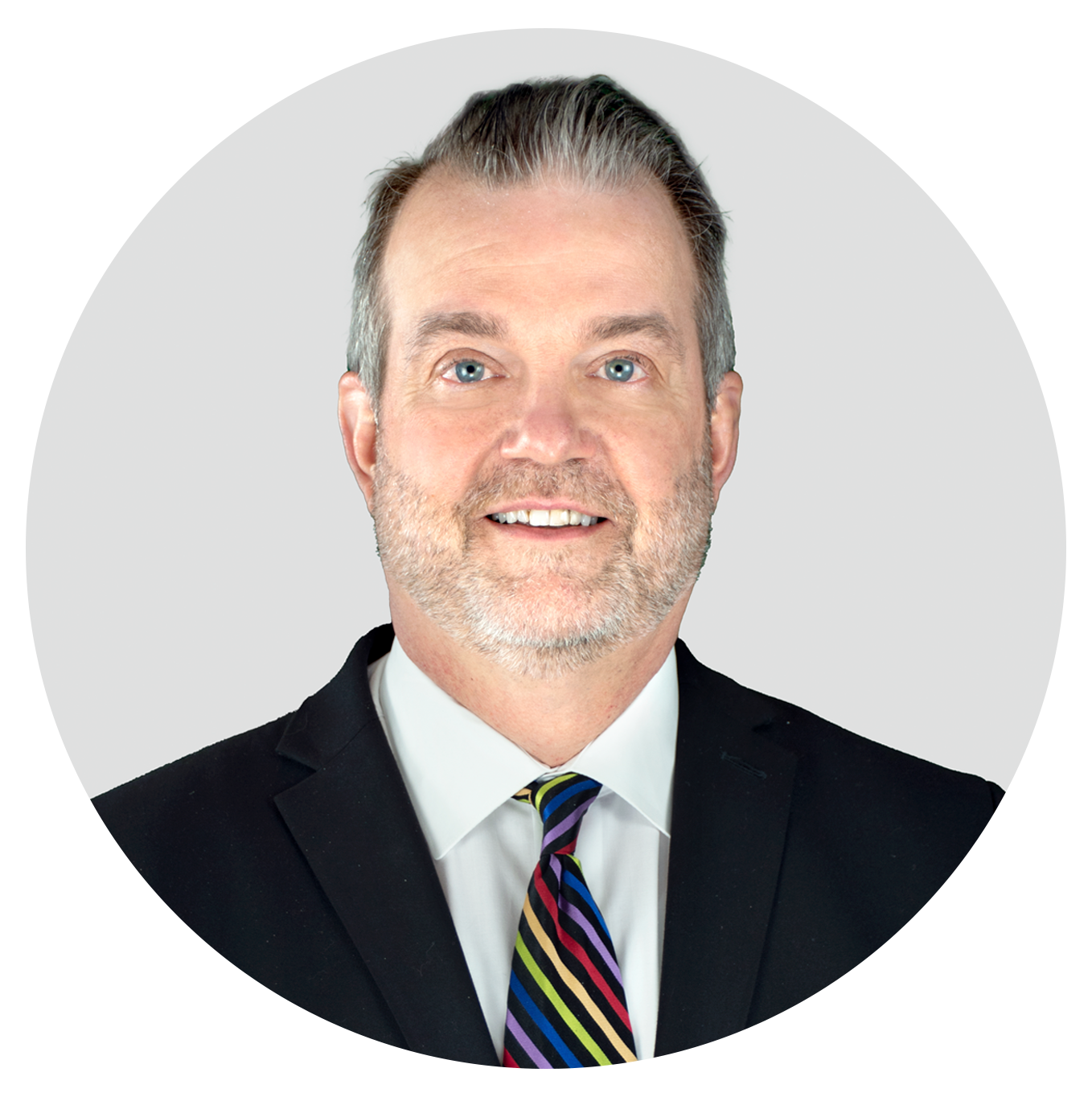 A middle-aged man with light skin, gray hair, and a beard, wearing a black suit, white shirt, and a colorful striped tie, smiling at the camera against a light gray background.