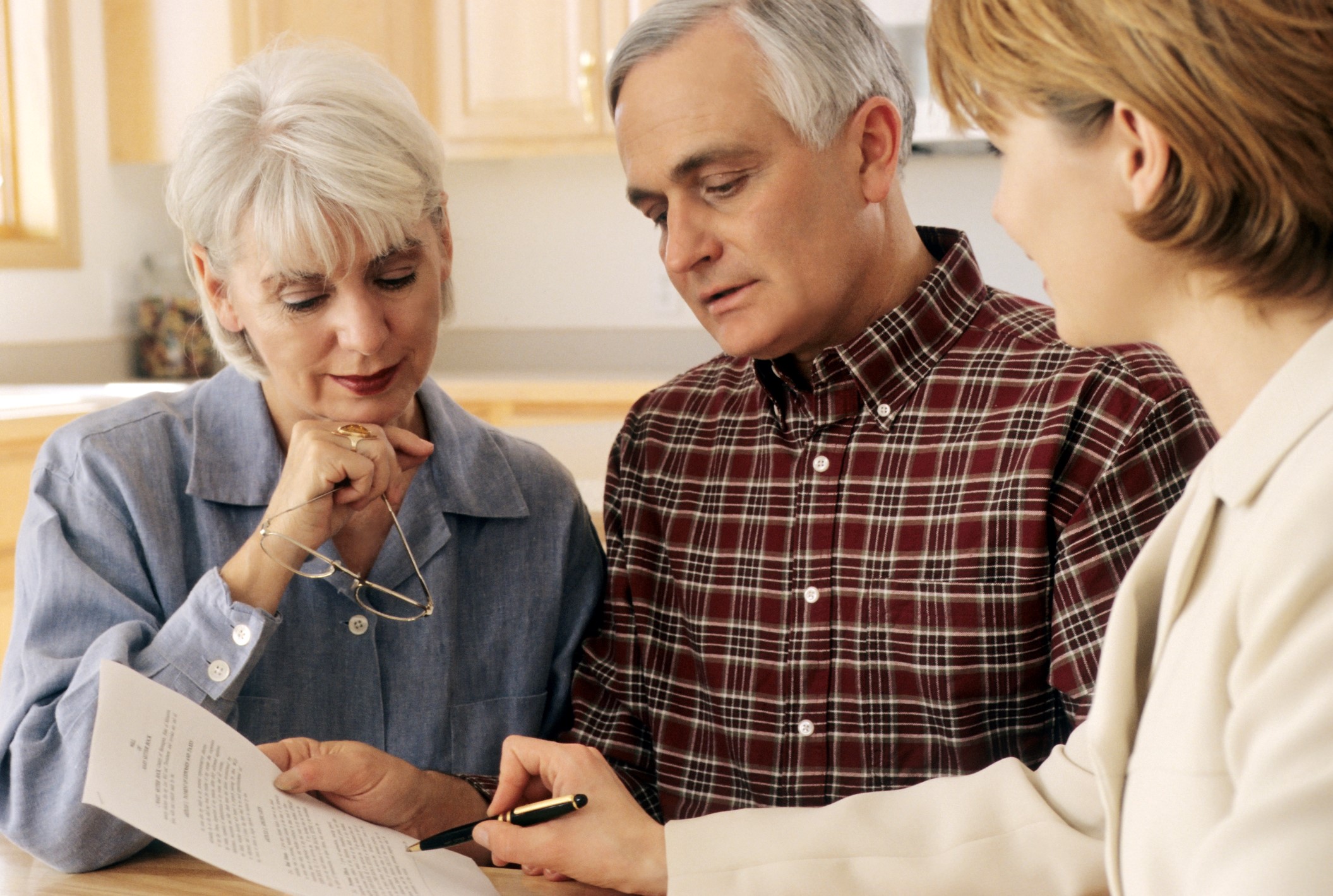 An older couple sits at a table reviewing a document with a professional woman, who is pointing to the paper, in a well-lit kitchen setting. The atmosphere appears focused and collaborative.