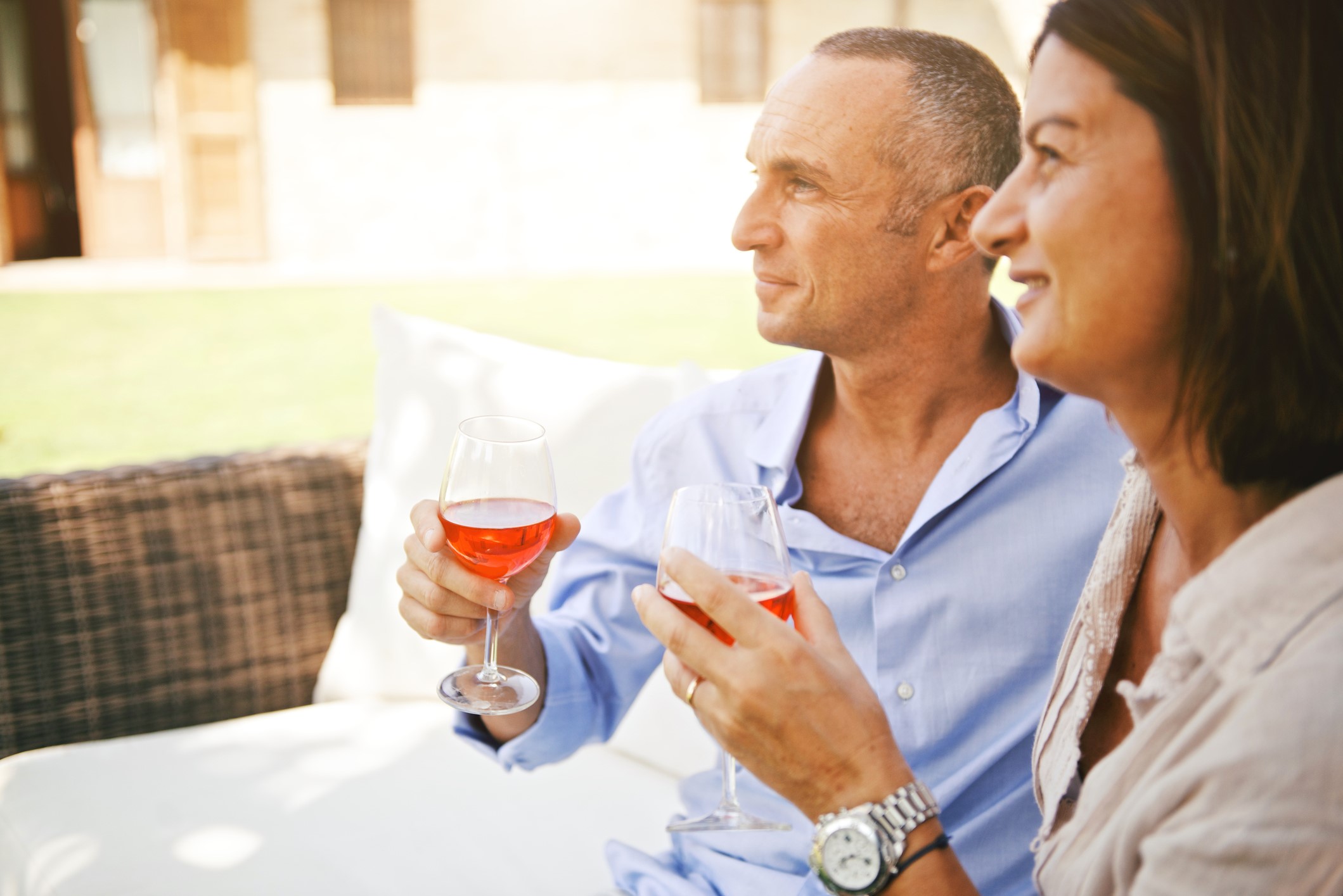 A man and woman sit closely together outdoors on a sunny day, each holding a glass of rosé wine. They are smiling and looking off into the distance, appearing relaxed and content.