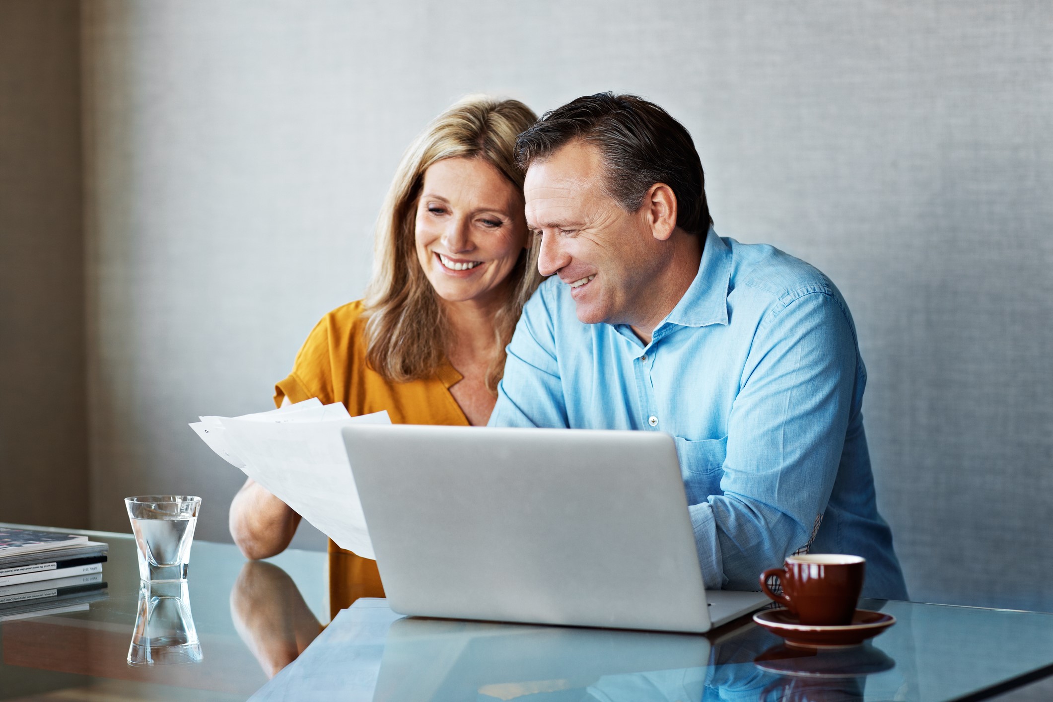 Smiling middle-aged couple sits at a glass table, looking at papers together in front of an open laptop, with a glass of water, a cup of coffee, and magazines beside them.
