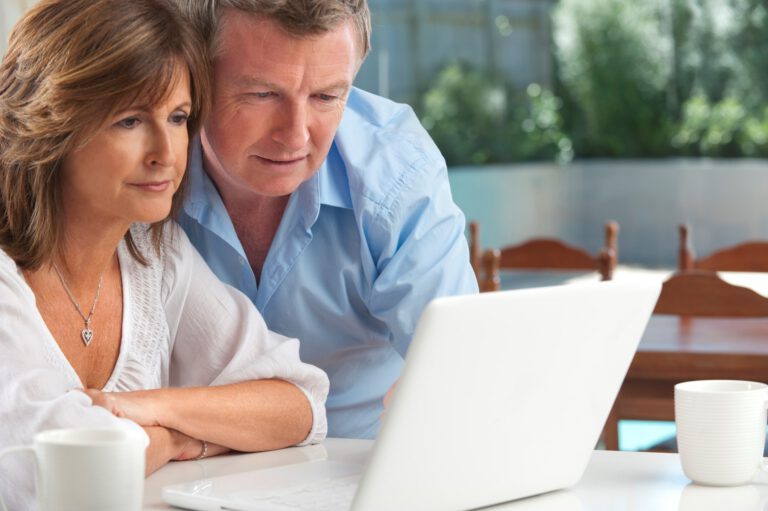 A middle-aged couple sits closely together at a table, looking intently at a laptop screen. They appear focused and engaged, with coffee cups on the table in a bright, homey setting.