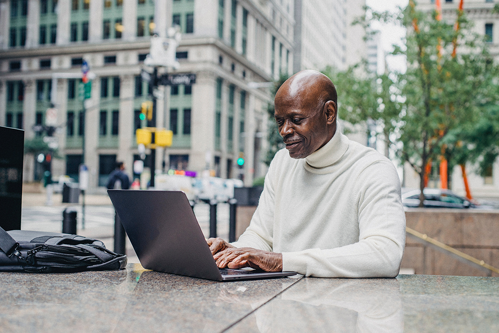 An older man wearing a white turtleneck sweater sits outdoors at a stone table, using a laptop and smiling, with city buildings and trees in the background.