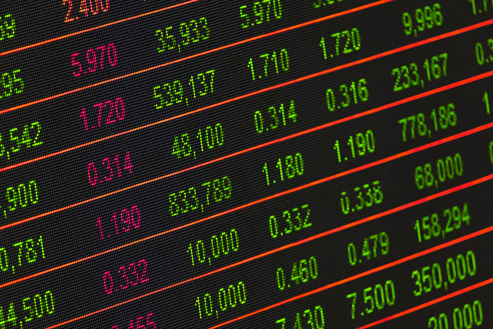 A digital display shows rows of stock market data with numbers in green, pink, and white on a black background, representing prices, volumes, and financial figures.