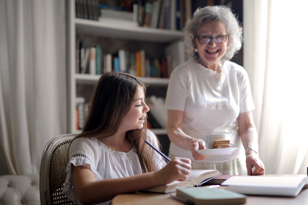 An elderly woman smiles while bringing a plate of cookies to a young girl who is writing in a notebook at a desk, with bookshelves in the background.