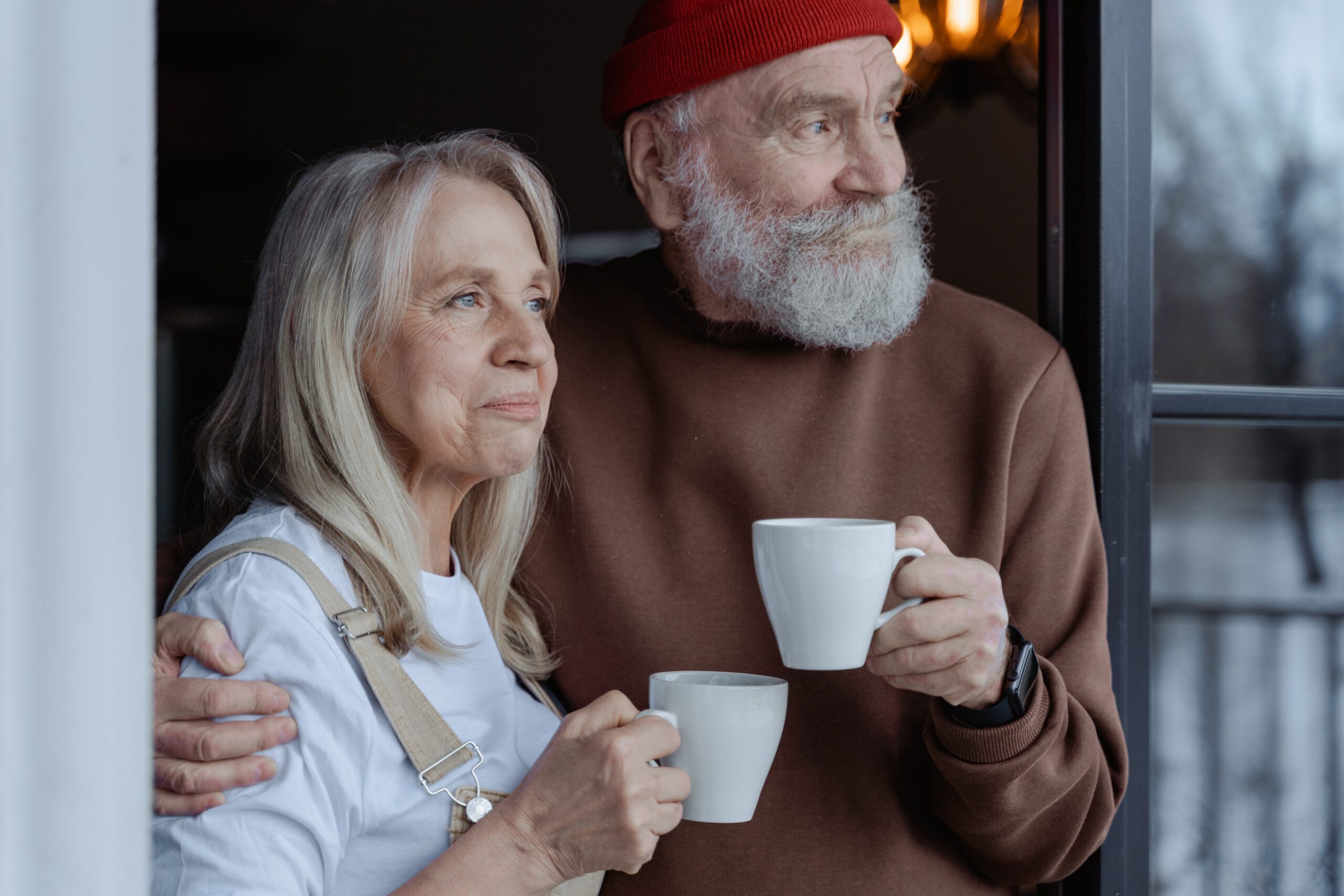 An older woman and man stand together by a window, smiling and holding white mugs. The man wears a red beanie and brown sweater, while the woman wears a white shirt and overalls. They appear relaxed and content.