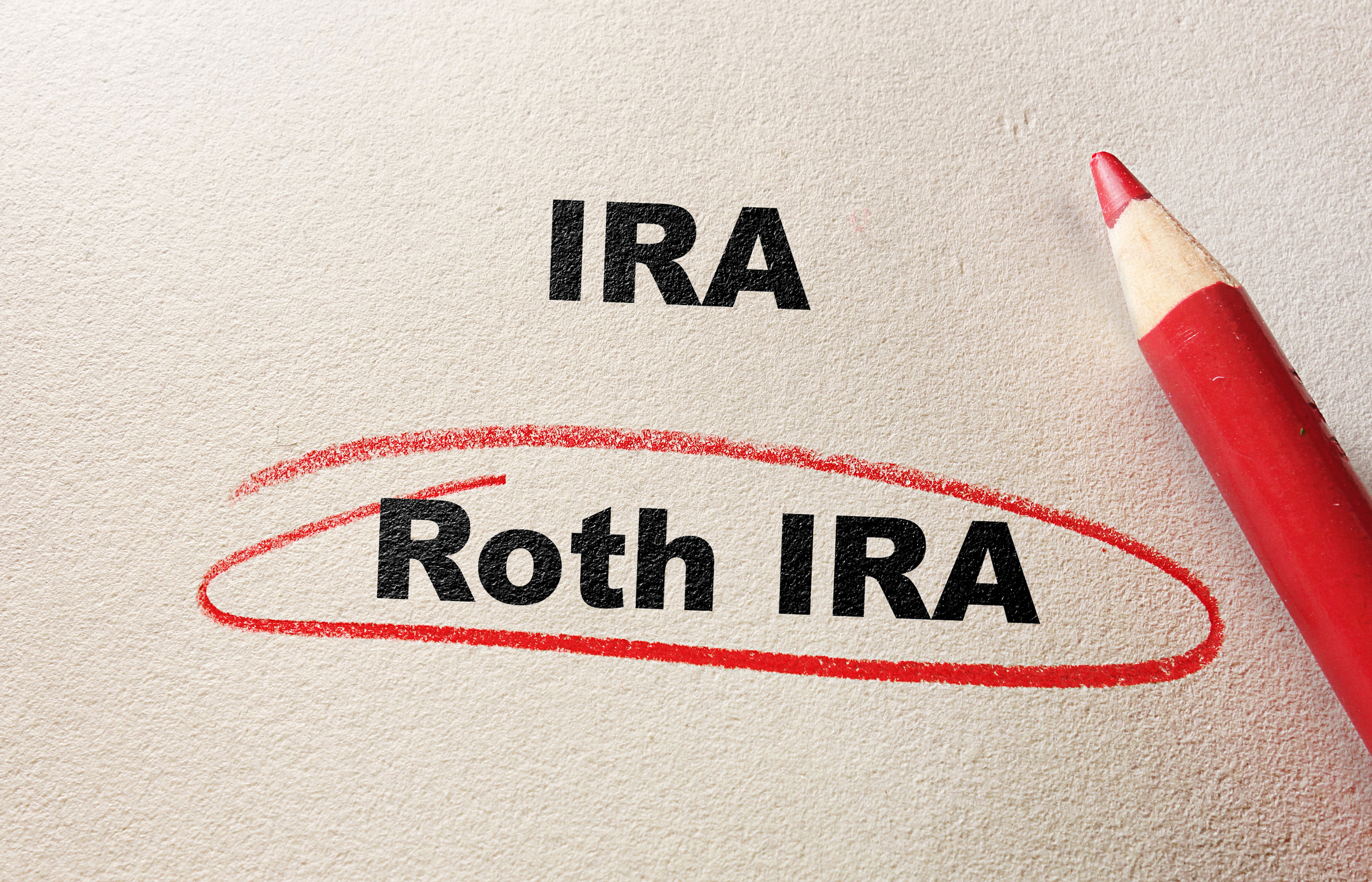 A red pencil underlines the words “Roth IRA” in red, below the word “IRA” written in black on a textured, beige surface.