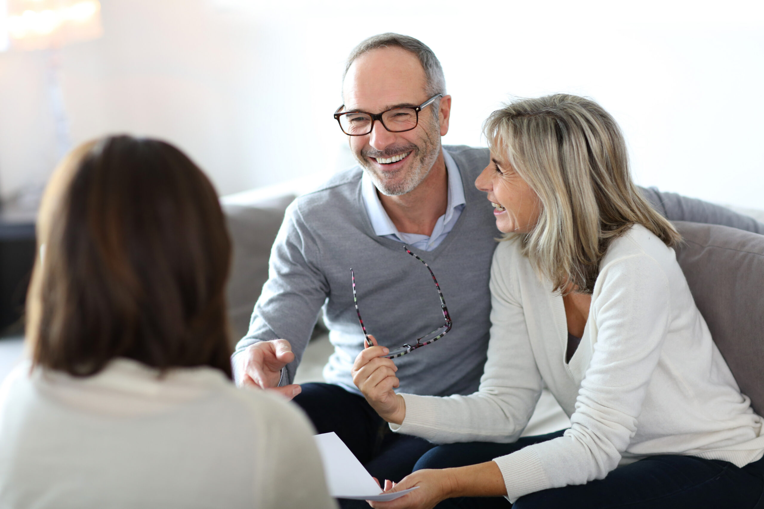 A smiling mature couple sits on a couch, talking to a woman with her back to the camera. The man holds glasses, and both appear engaged and happy during the conversation, possibly a consultation or meeting.