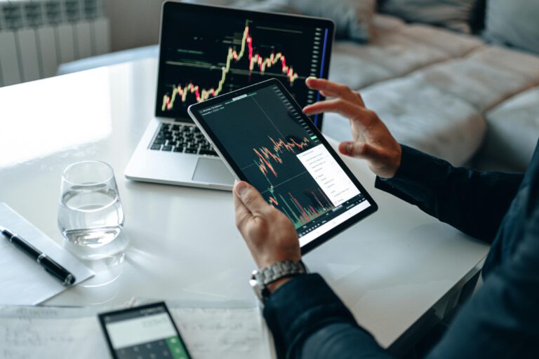 Person analyzing stock market charts on a tablet, with a laptop displaying graphs in the background, a glass of water, and financial documents on a white desk.