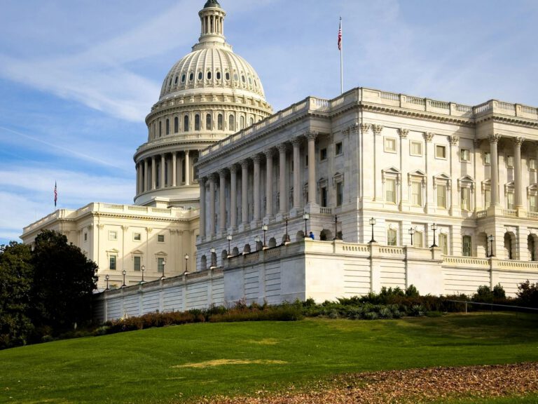 The U.S. Capitol building with its large dome, white columns, and American flag, stands behind a grassy lawn under a blue sky with wispy clouds.