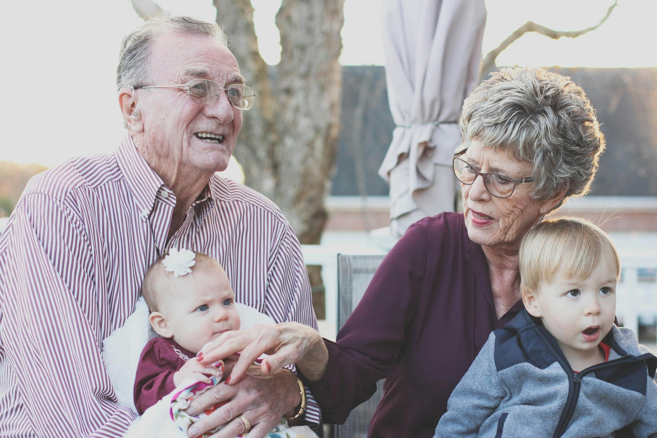 An elderly man and woman sit closely together outdoors, holding a baby and a young child on their laps. All are smiling or looking engaged, enjoying a pleasant moment together.